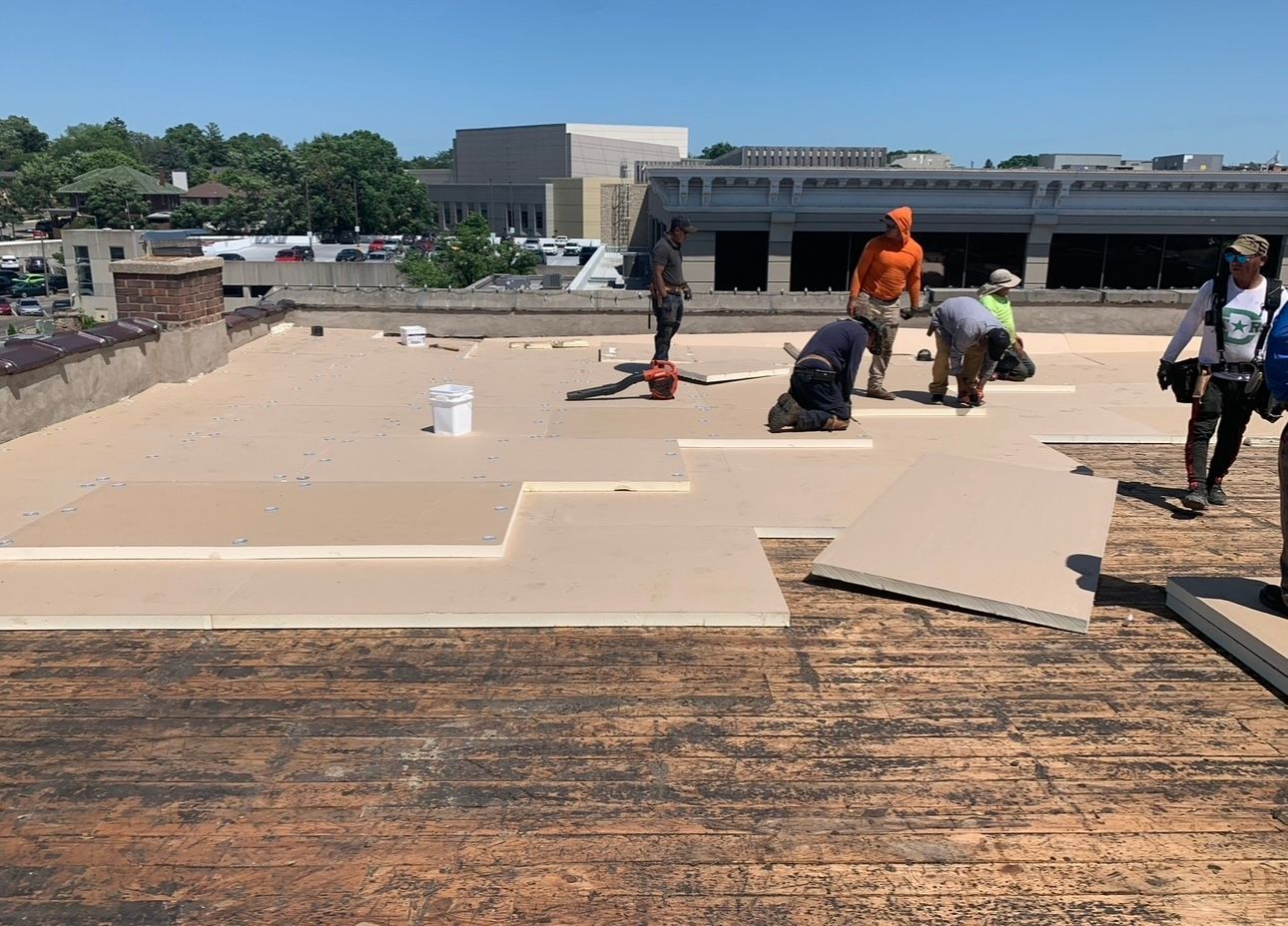 Workers installing insulation panels on a flat rooftop under a bright, sunny sky. Beige panels are being fitted, with some workers kneeling.