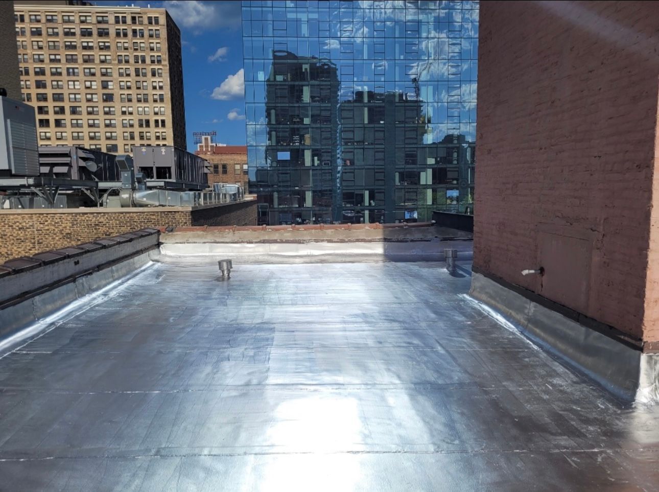 Rooftop view with a reflective modern building and other city structures in the background. The roof has silver sheeting.