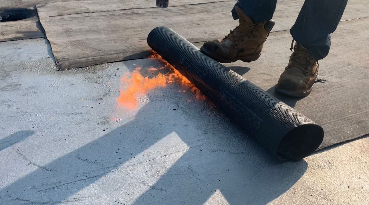 A worker rolls out roofing material and uses a torch to adhere it to a rooftop. Fire is visible.