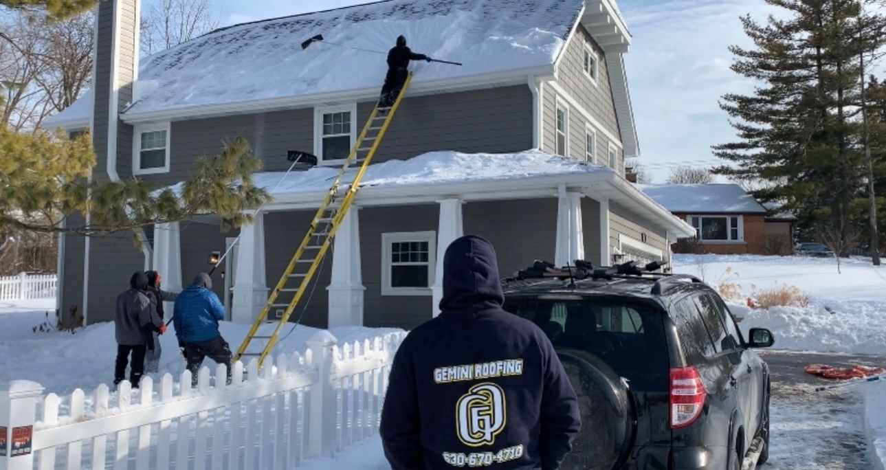 People clearing snow from a house roof with a ladder, car parked in driveway. It is winter.