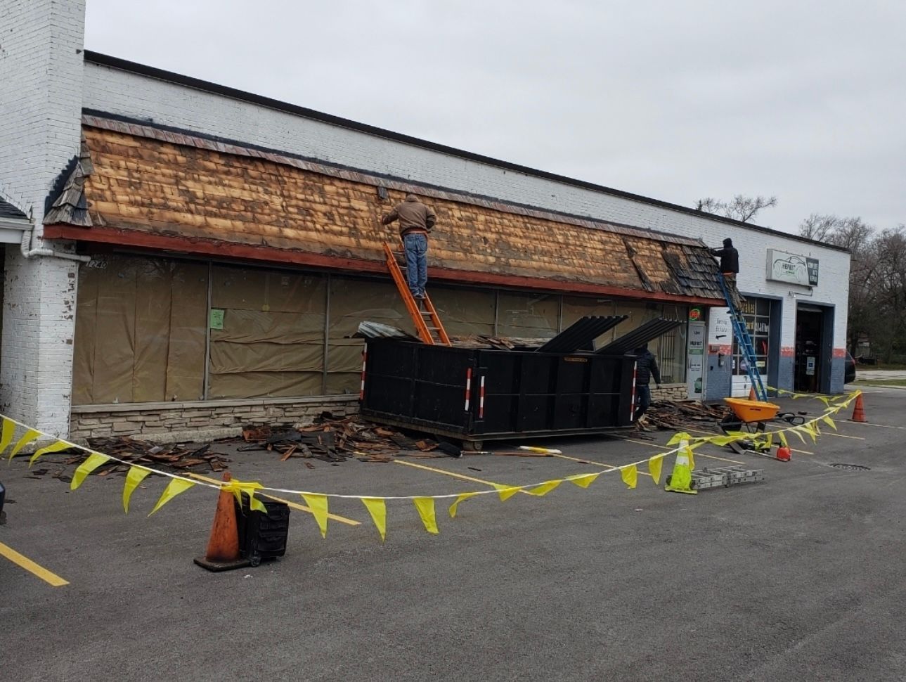 Construction worker on a ladder removing a building's awning. A large dumpster sits below, with safety tape and cones in the parking lot.