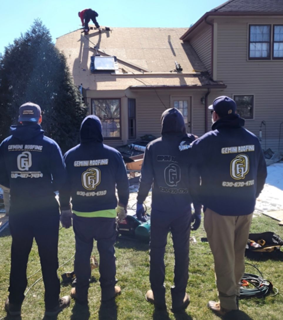 Four roofers in matching sweatshirts stand in a yard, looking at a rooftop where another roofer is working.