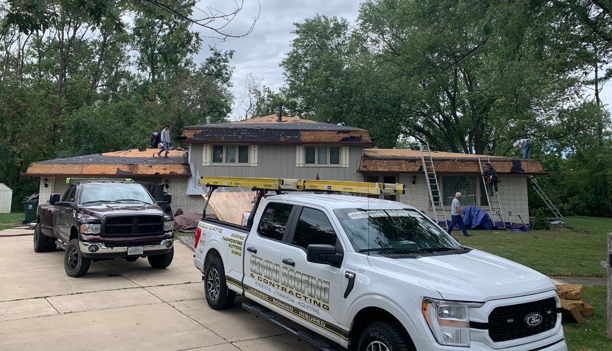 Roofers working on a house; two trucks are parked in front of the house on a driveway. Green trees are in the background.