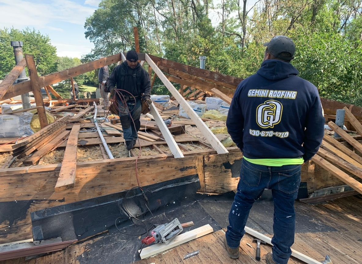 Two roofers work on a partially dismantled roof. One carries tools, the other faces away, wearing a navy hoodie.