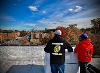Two roofers on a rooftop, looking at a cityscape under a blue sky with autumn foliage. One wears a black jacket, the other an orange hoodie.