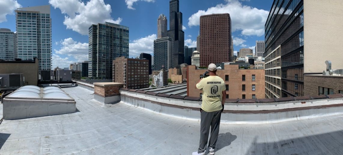 A person stands on a rooftop overlooking a cityscape with tall buildings under a blue sky.
