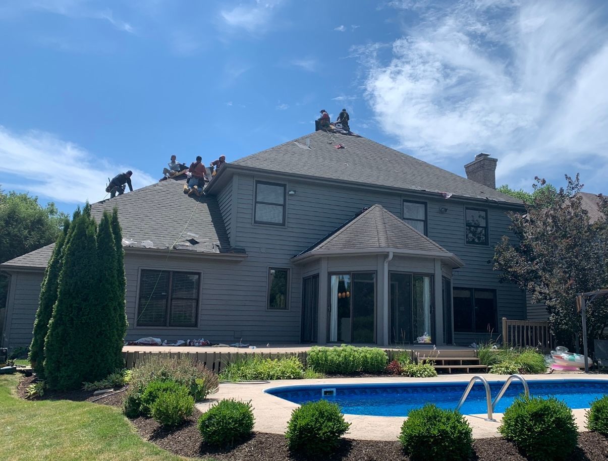 Workers replace roof shingles on a two-story gray house with a pool in the backyard on a sunny day.