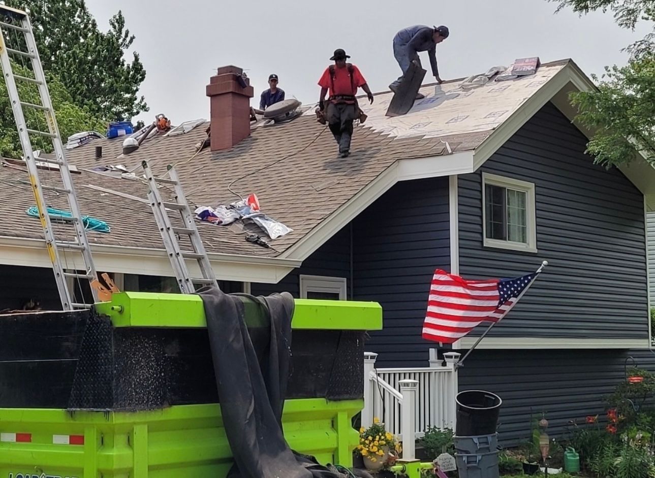 Roofing crew working on a house with gray siding and an American flag. Green dump truck and ladders present.