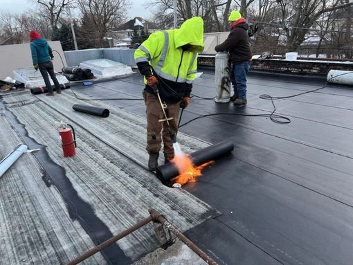 Workers on a flat roof use a torch to heat a black roofing membrane. One worker in yellow is in focus.