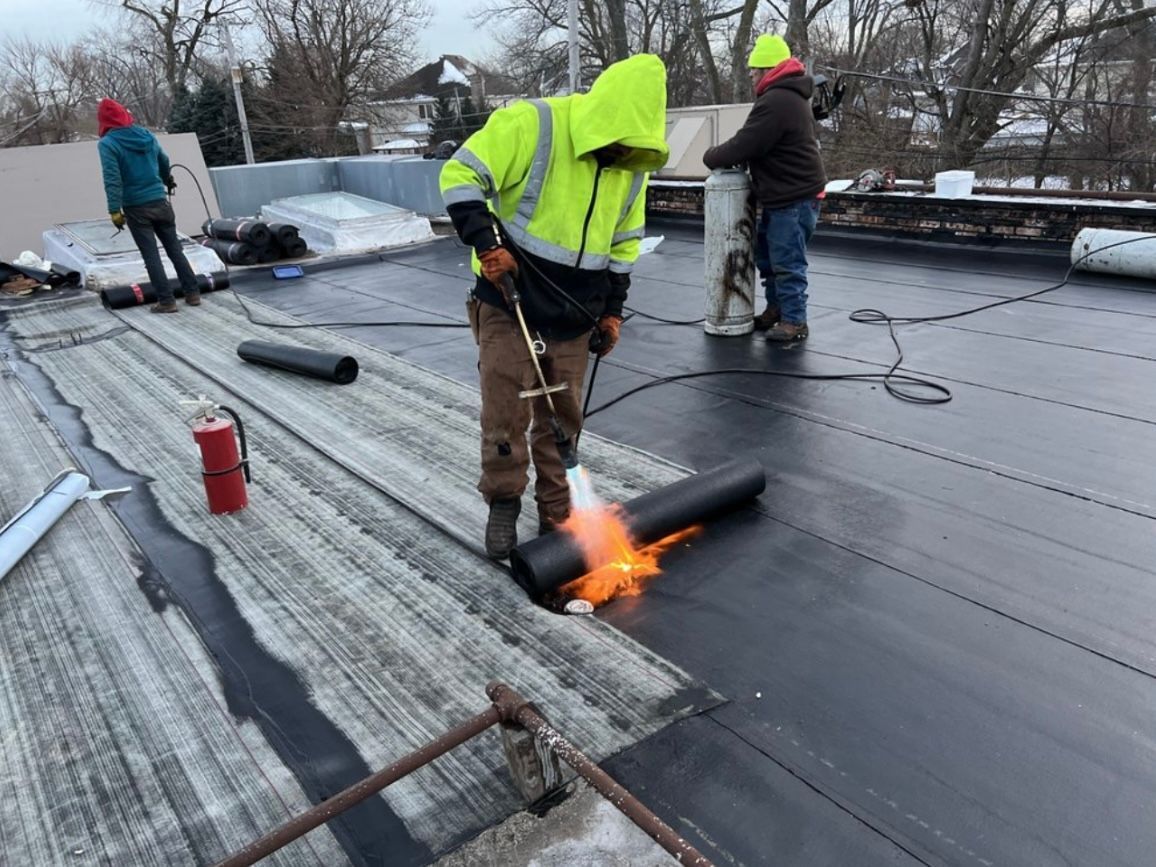 Construction workers torching a black roofing membrane on a rooftop. One worker in yellow safety gear is holding the torch.