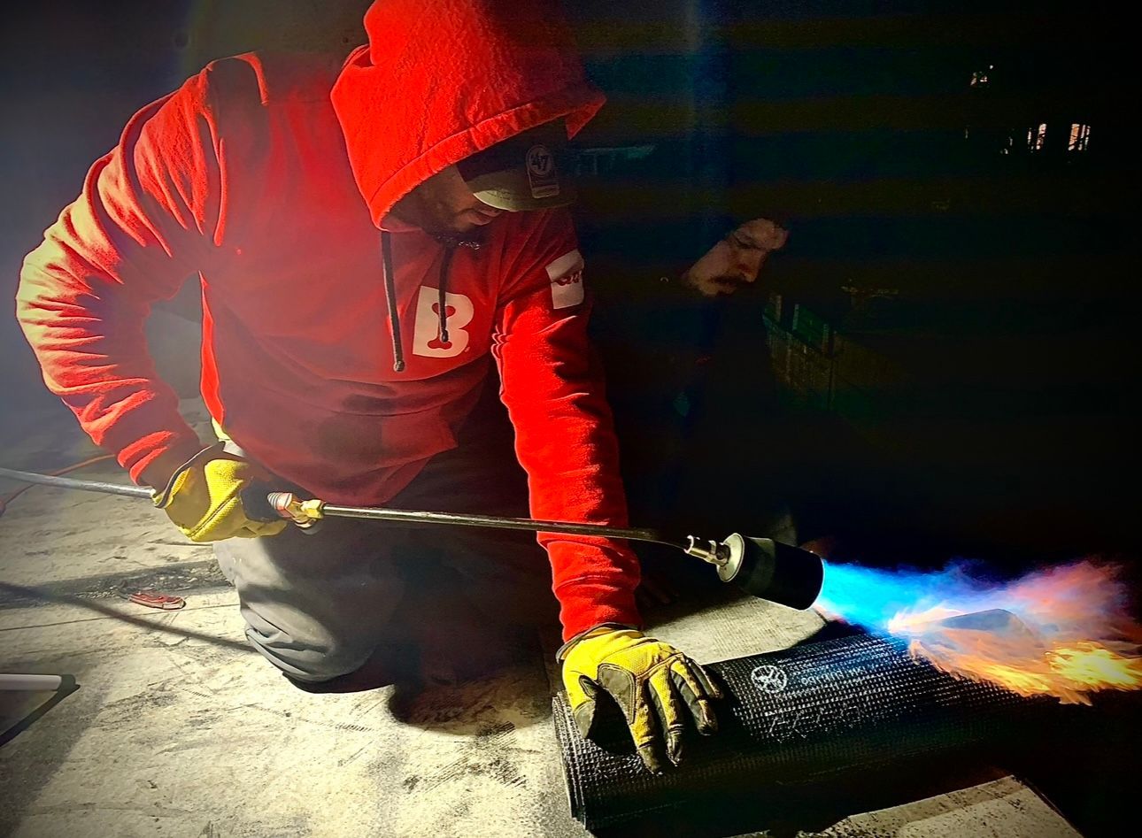 A person in a red hoodie and yellow gloves uses a blowtorch to apply something to a dark surface, possibly roofing, outdoors at night.