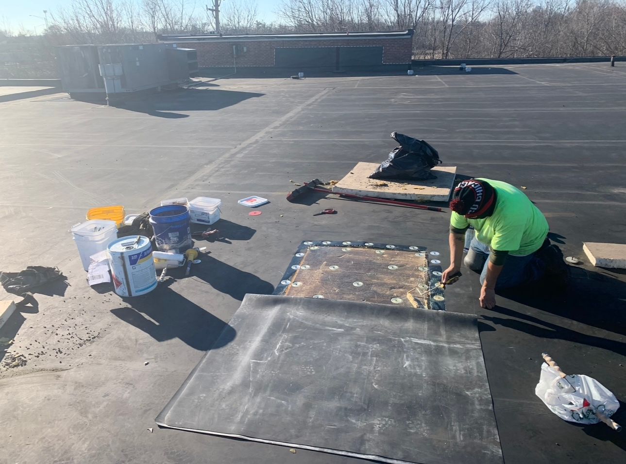 A person in a green shirt repairs a black flat roof on a sunny day. Tools and materials are scattered around the work area.