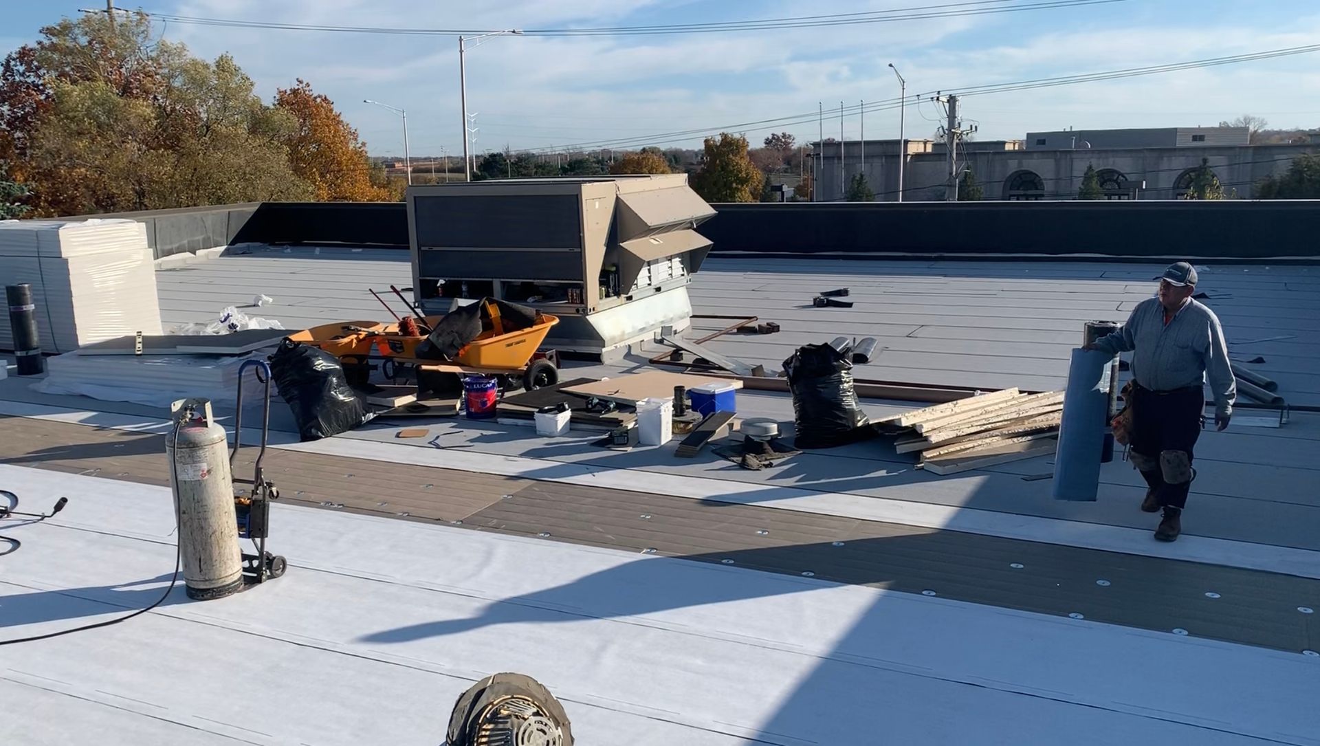 Workers on a commercial rooftop installing white roofing material. Equipment and supplies are scattered around the work area. A man walks toward the right side of the frame.