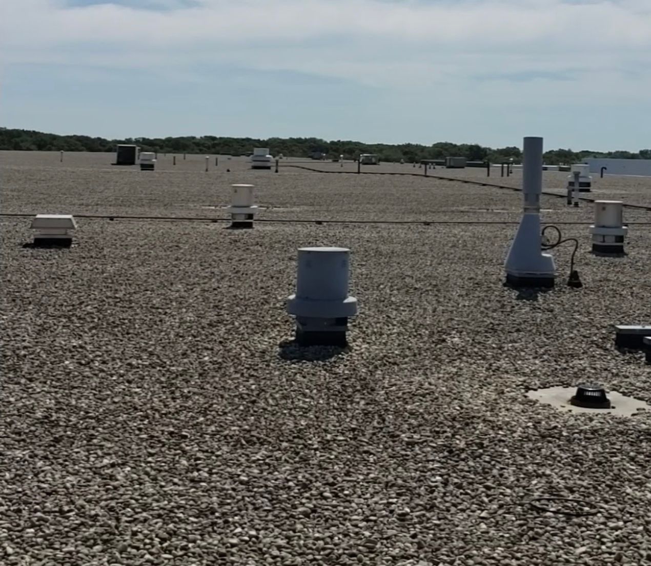 A flat, gravel-covered rooftop with various vents and pipes. The background shows a distant tree line and a blue sky.