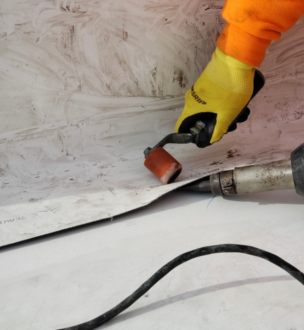 A worker uses a roller and heat gun to seal a white membrane. The setting appears to be an interior construction site.