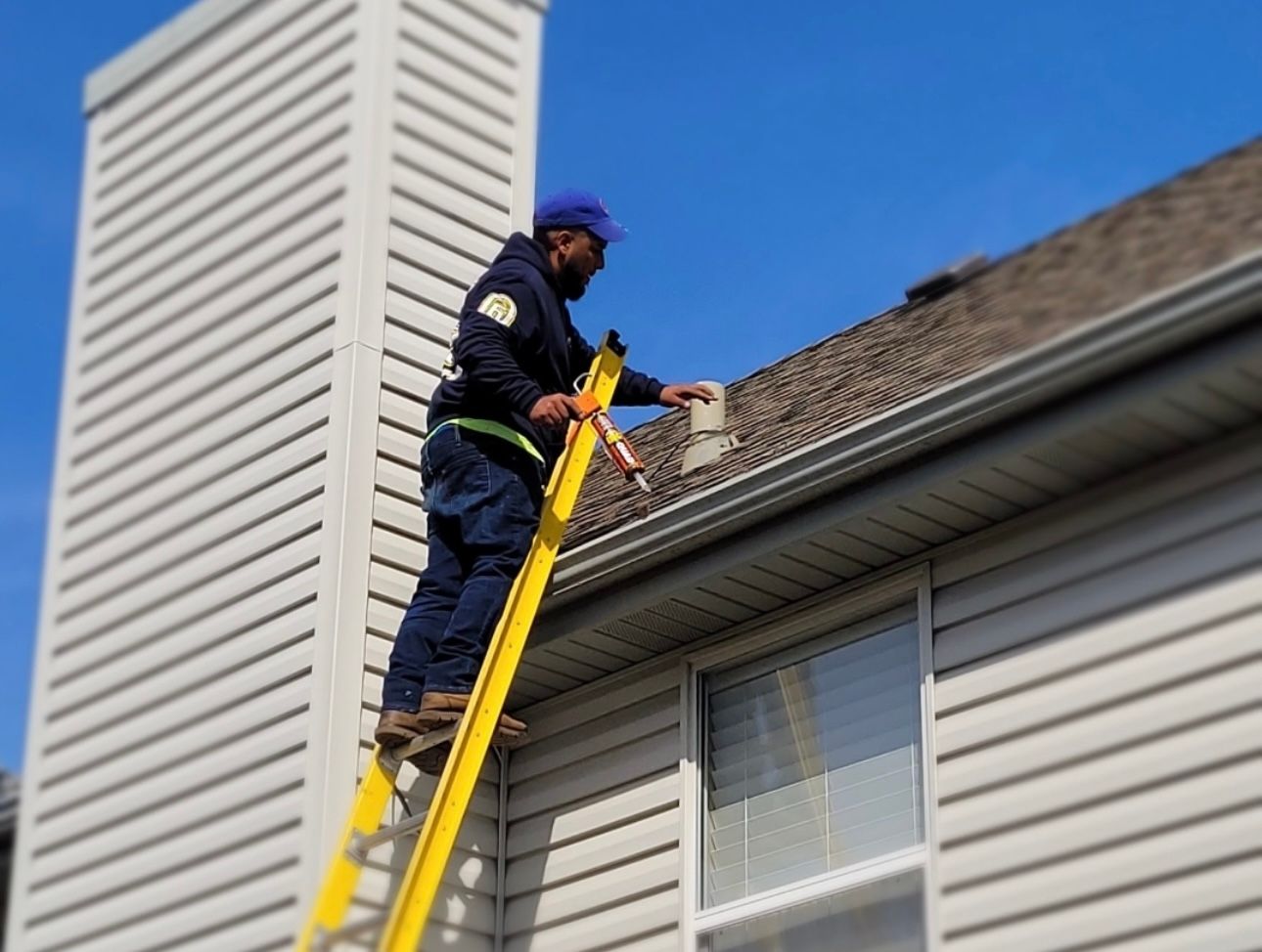 A man on a yellow ladder is caulking a roof next to a chimney. He wears a blue hat and jacket.