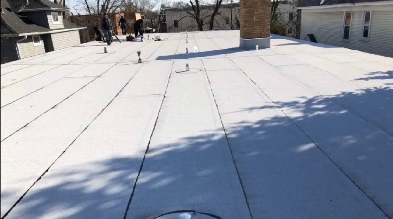White flat roof with dark seams, a chimney, and a few people in the background. Sunlight casts shadows on the roof.