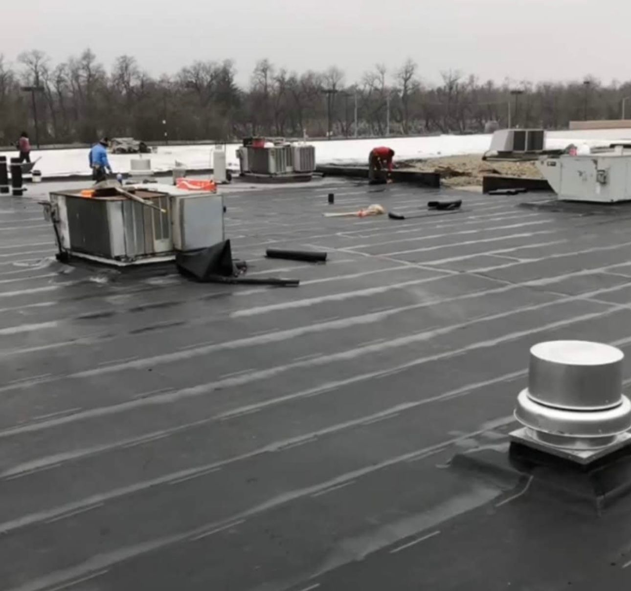 Workers repairing a flat, black commercial roof with HVAC units and vents. Overcast day with trees in the background.
