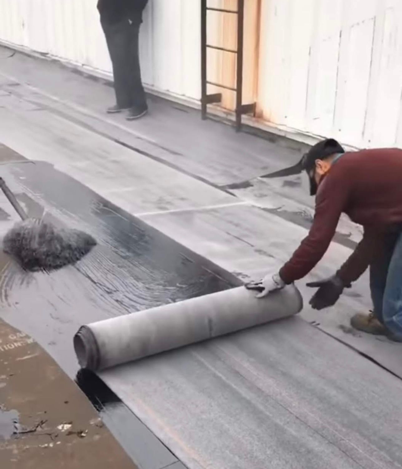 Two workers installing roofing material on a flat roof. One rolls out a gray sheet, while another applies a black sealant with a brush.