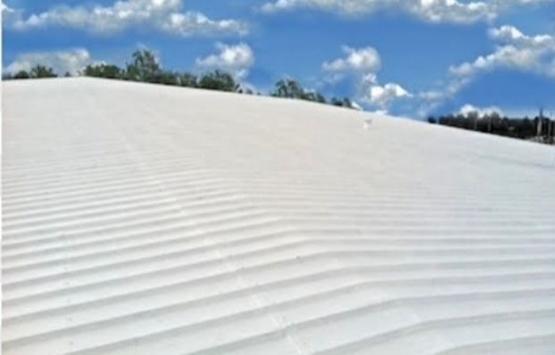 White, corrugated roof sloping upwards toward trees and a blue sky with clouds.