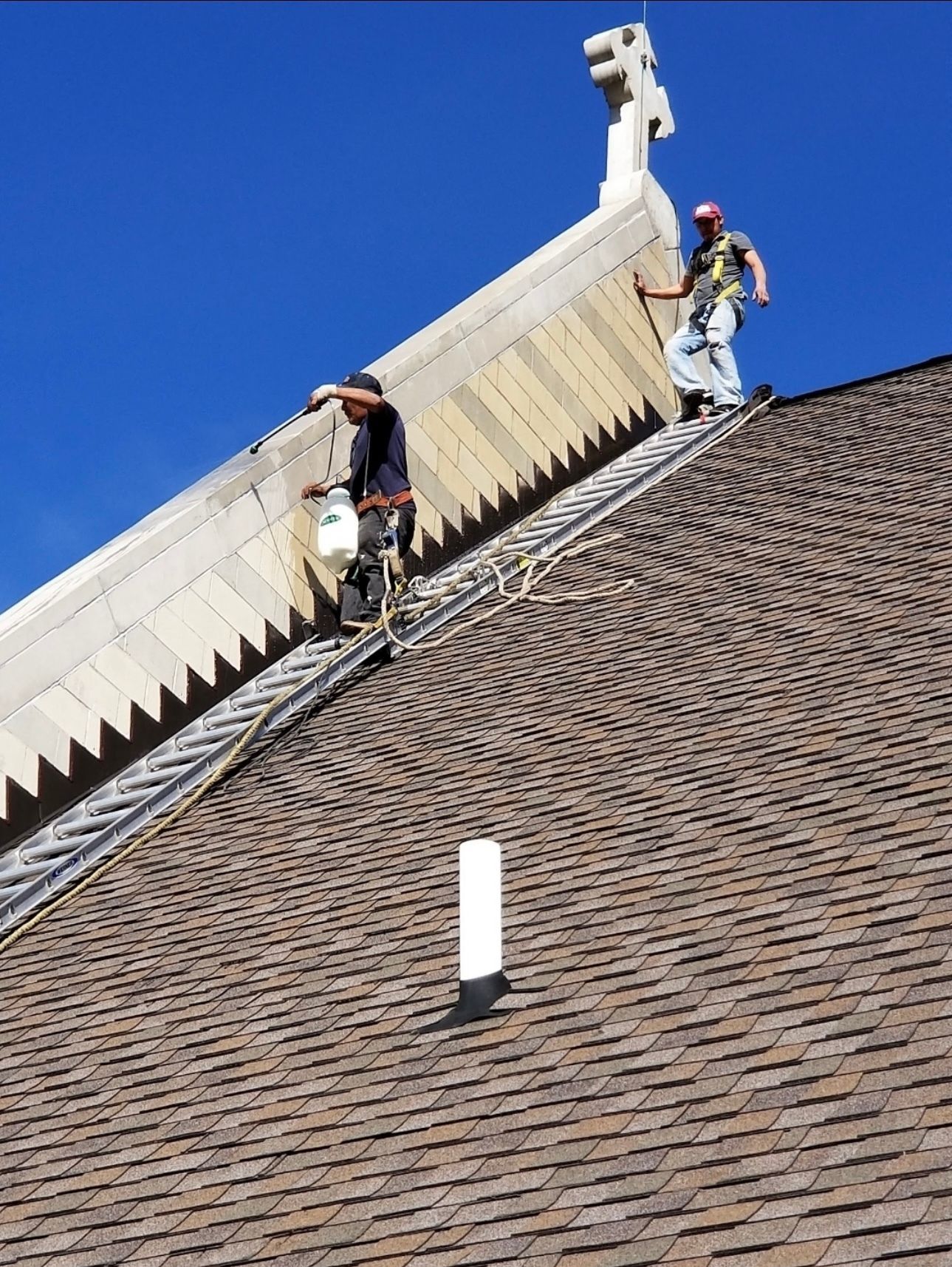 Two workers on a steeply sloped roof near a decorative peak. One paints, the other is secured with a harness. Sunny, blue sky.
