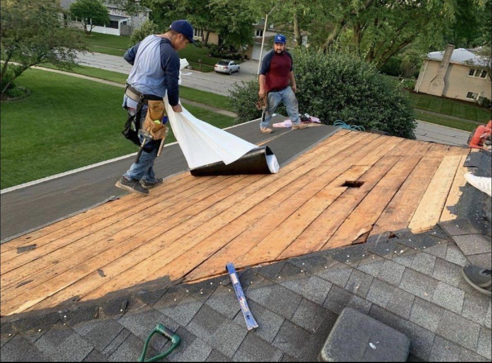 Two roofers installing underlayment on a partially shingled roof. One unrolls the black underlayment, while the other stands nearby.