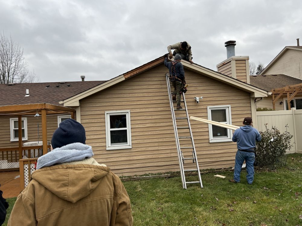 Construction workers replacing roof shingles on a tan-sided house, one on a ladder, another on the roof. Two other workers assist on the ground. Overcast day.