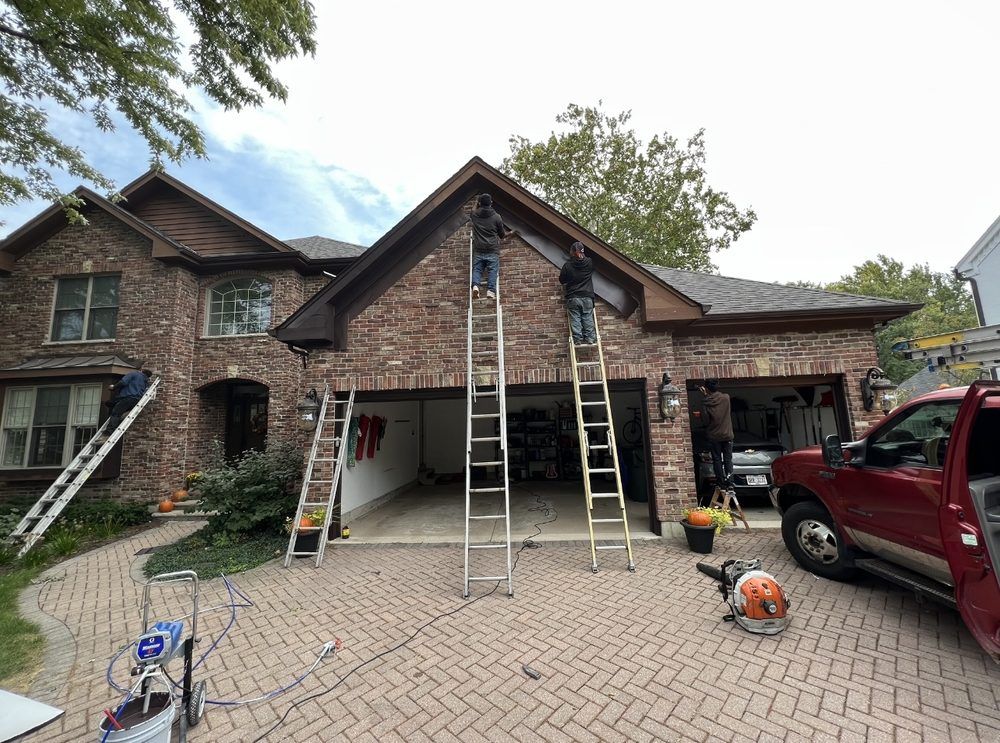 Two workers on ladders repairing the brown brick house roof over a garage. A truck and leaf blower are in the driveway.