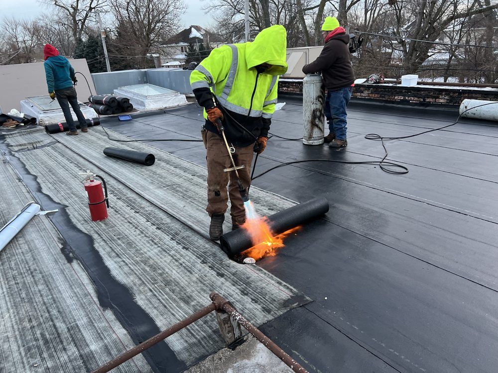 Roofers installing a black membrane roof. One worker in a yellow safety jacket uses a torch to seal the roofing material. Others are nearby.