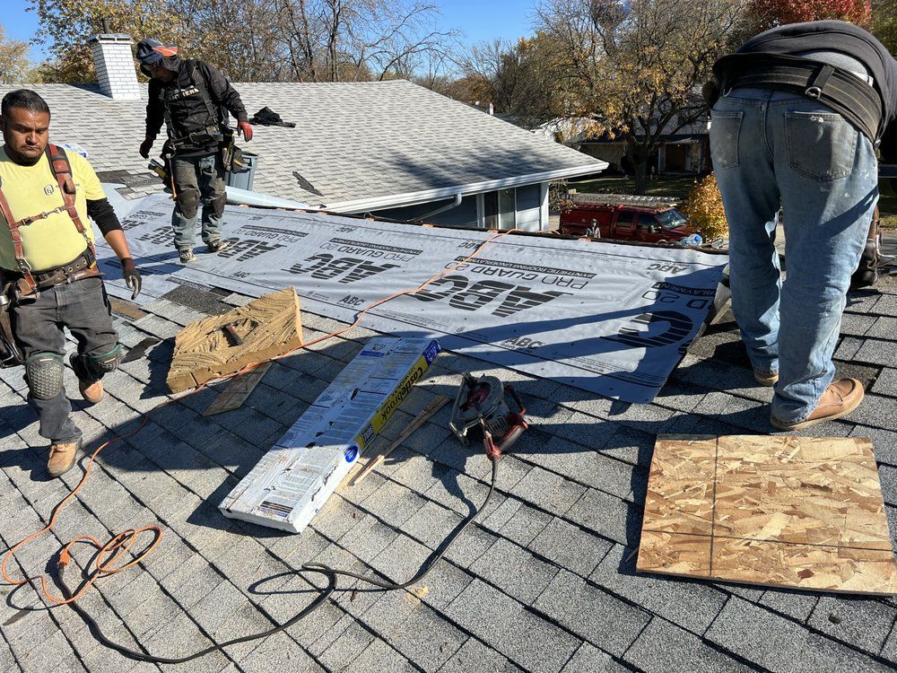 Roofing workers installing new shingles on a residential roof, applying underlayment. Blue sky in the background.