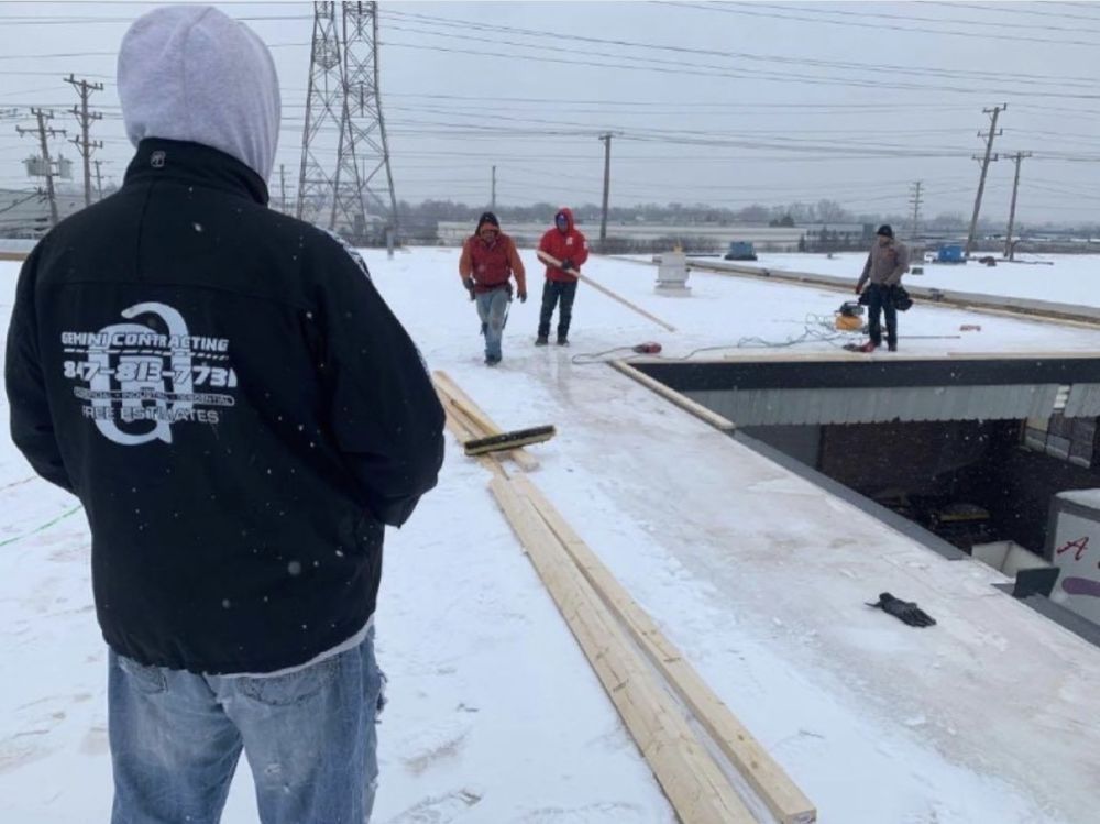 Construction workers on a snowy rooftop. A man in a hooded jacket looks at the team. Other workers are walking and working on the flat roof.