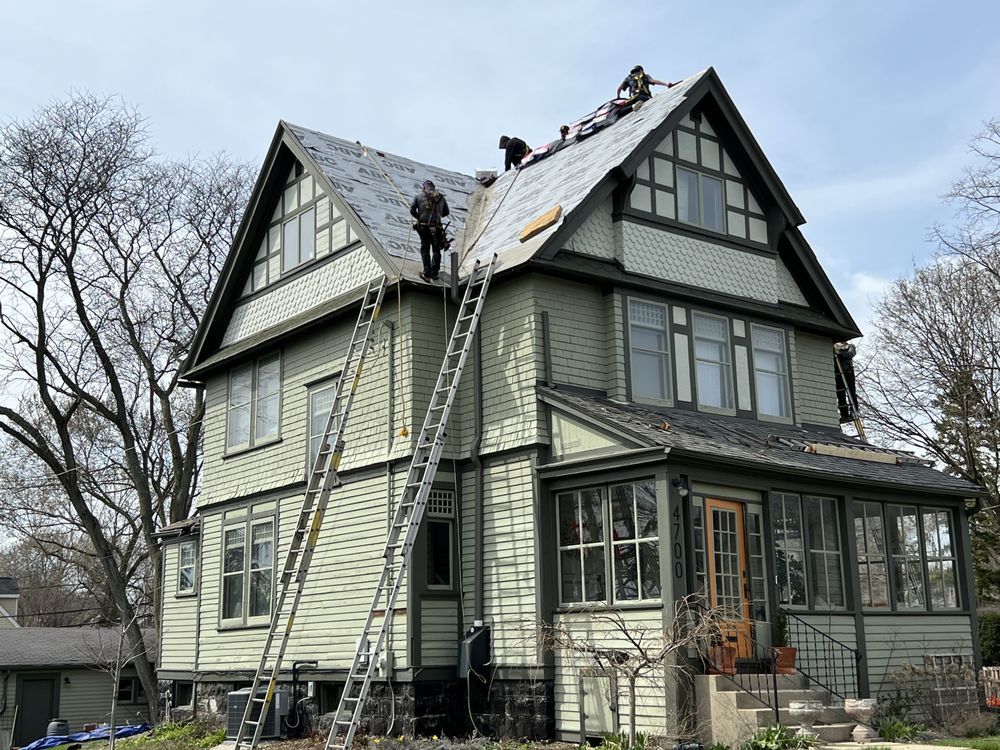 Roofers replacing shingles on a three-story light green house with dark green trim. Ladders lean against the house.