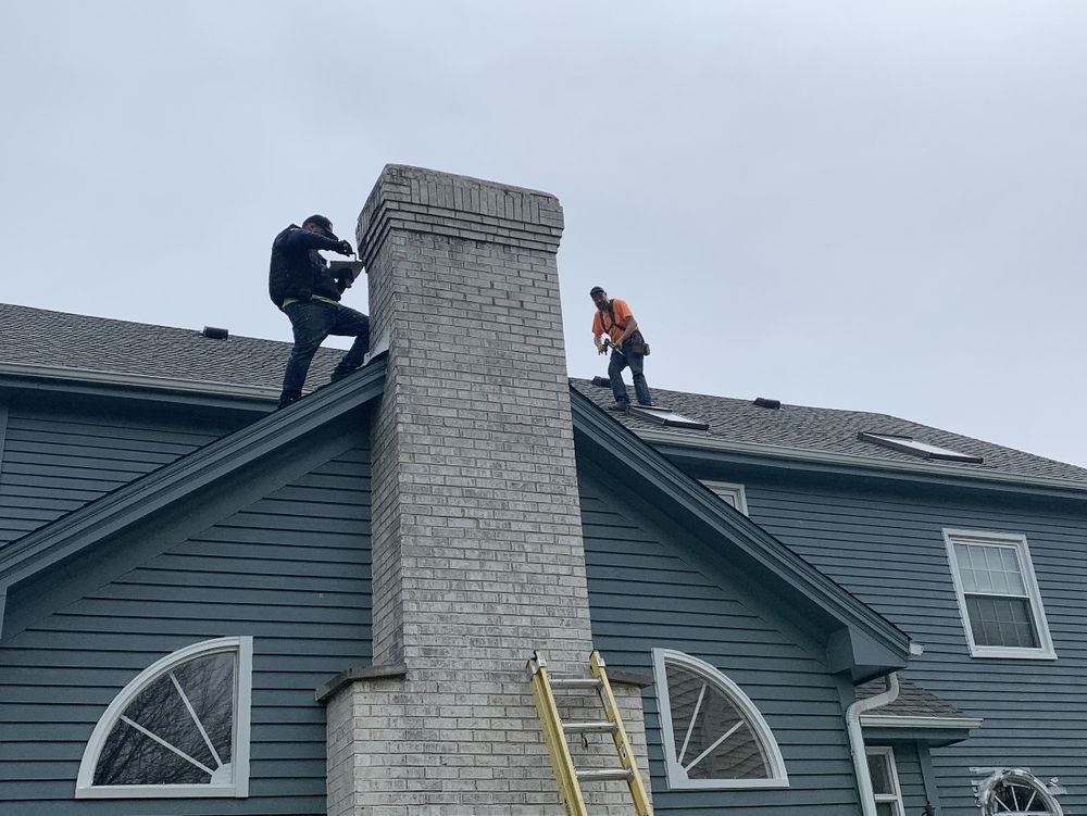 Two workers on a roof, repairing a brick chimney on a house with blue siding. One is using a tool on the chimney, the other is further down the roof.