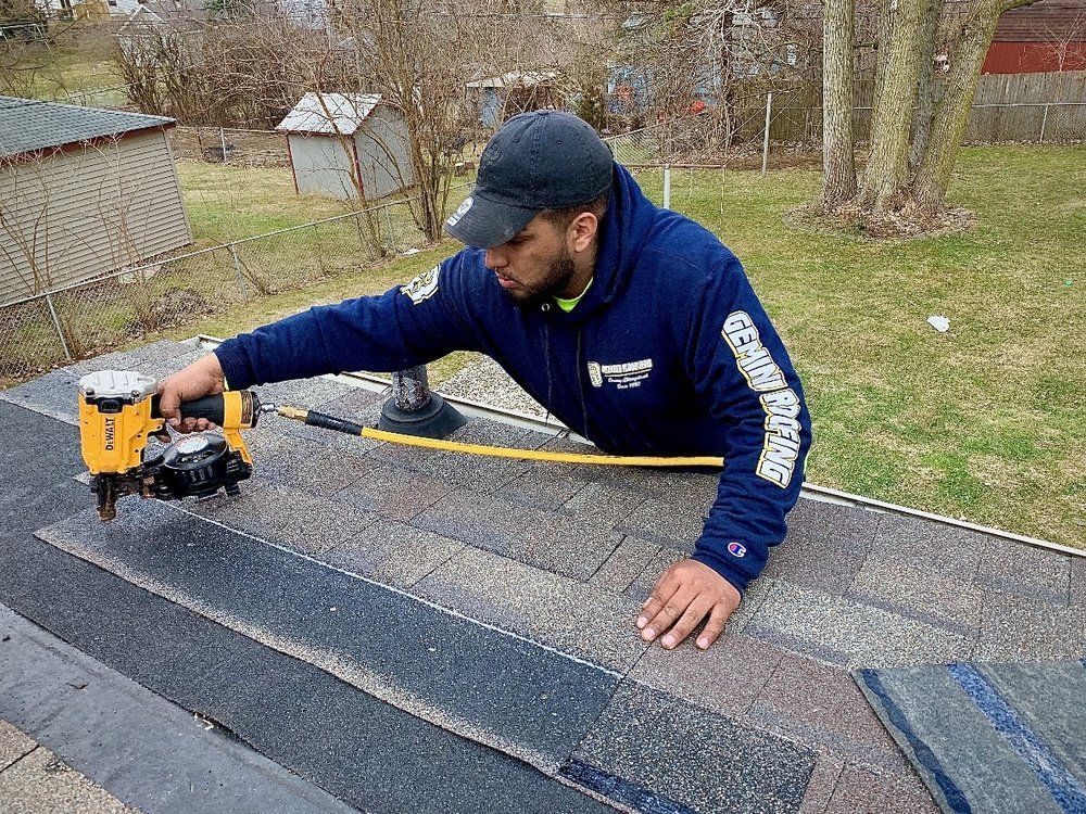 Roofer in a blue hoodie, wearing a hat, uses a nail gun to install shingles on a roof.