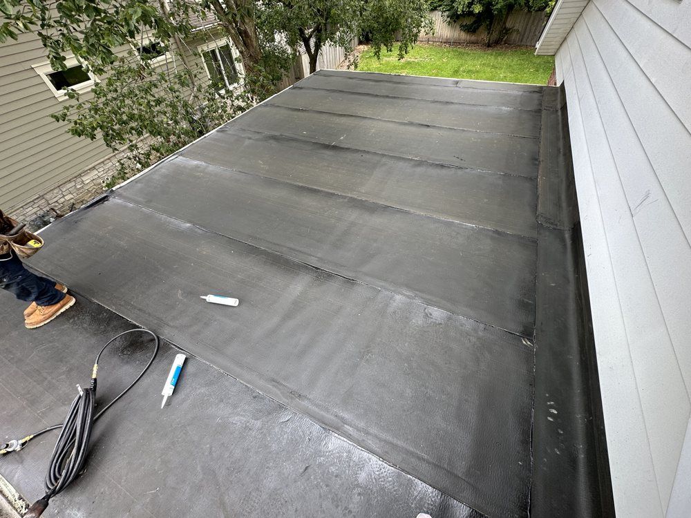 Flat black roofing with multiple seams; a worker in brown boots is visible on the left. The roof is against a white wall.