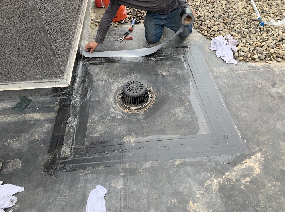 Person sealing a flat roof drain with gray tape. The roof is black, and there are tools and gravel nearby.