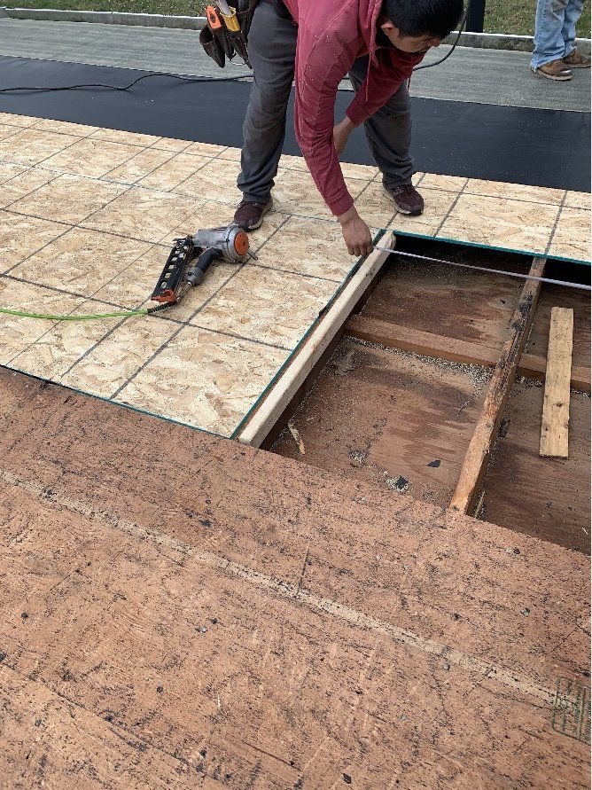 Person installing wood planks over a structural opening on a construction site. The person wears work clothes, and tools are nearby.