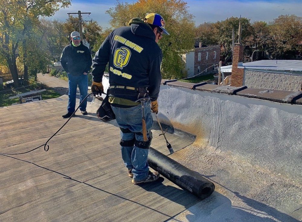 Two roofers installing roofing material on a flat roof. One uses a torch, the other watches. Sunny day.