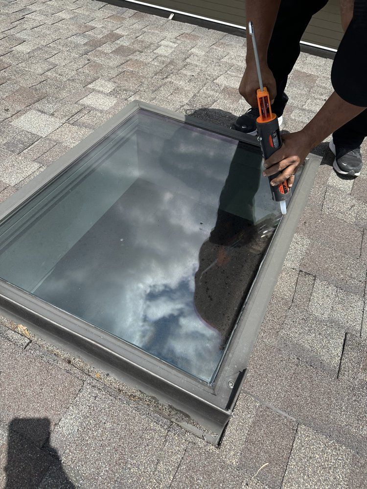 Person caulking around a rectangular skylight on a shingled roof, with the sky reflected in the glass.