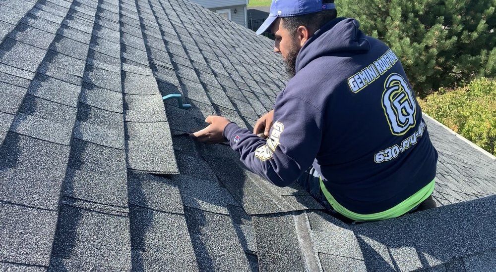 Roofer in a blue hoodie and hat repairs shingles on a gray rooftop on a sunny day.