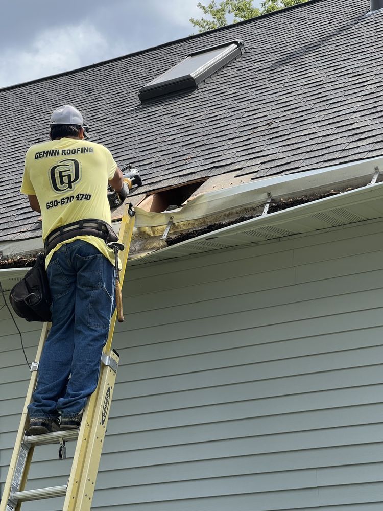 A construction worker on a ladder repairs damaged guttering on a house. He wears a yellow shirt and blue jeans.