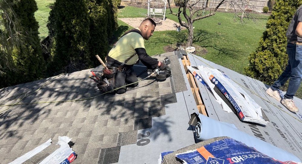 A roofer in a yellow shirt installs shingles on a rooftop. Tools and materials surround him, another person stands nearby.