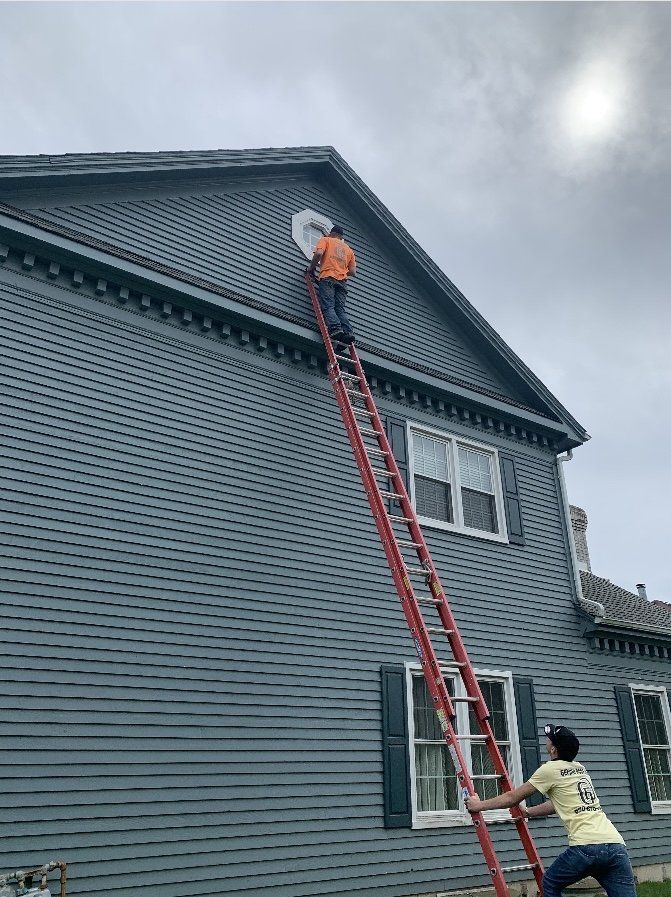 Two people on a ladder working on a gray house. One person is at the top near a round window. The other is stabilizing the ladder. Overcast sky.