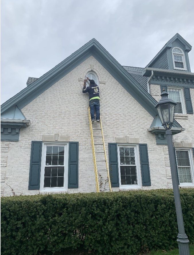 A person on a yellow ladder works on a round window high on a white brick house with teal trim.