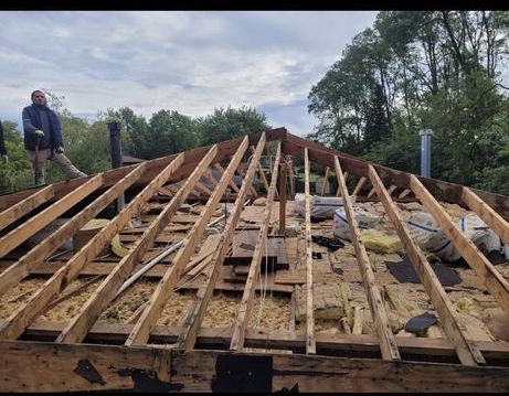 Person on a roof with exposed wooden rafters, trees and cloudy sky in the background. Roof construction or repair in progress.