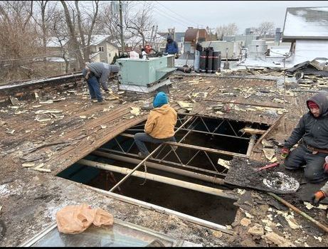 Workers on a rooftop repair an open skylight. One person sits inside, others work around the opening in a cloudy, outdoor setting.