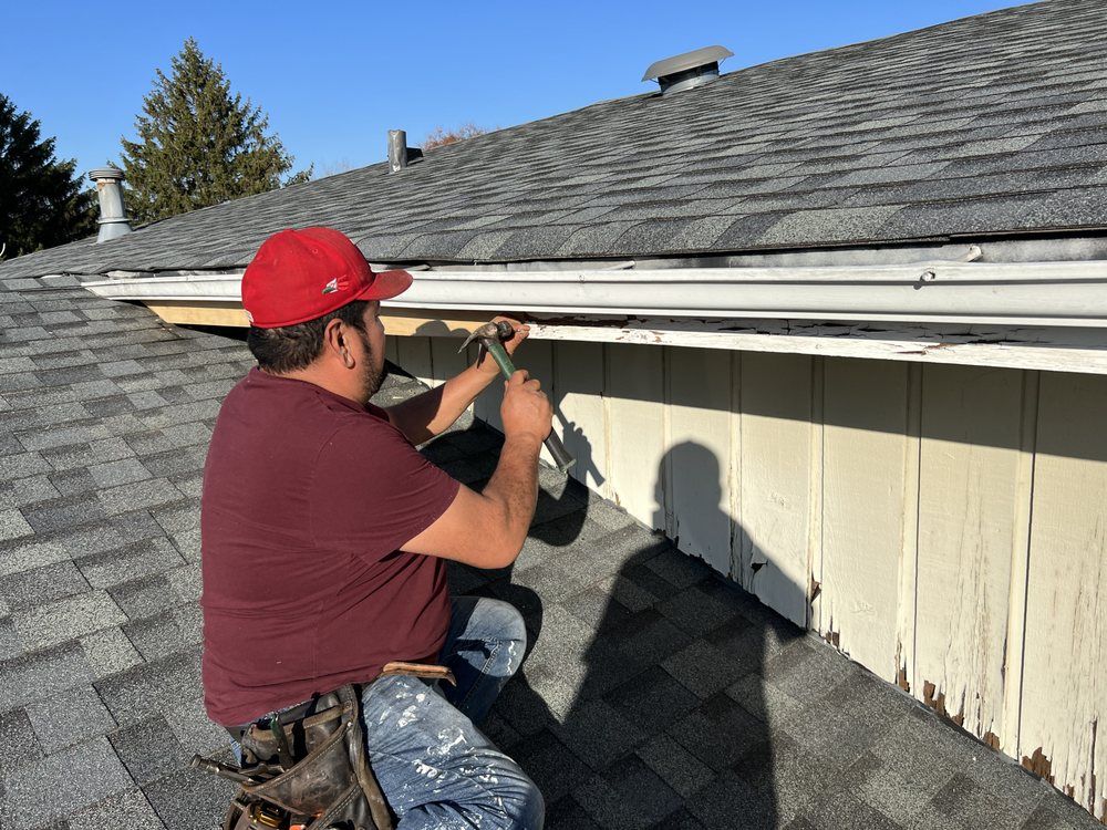 A person wearing a red hat repairs gutters on a rooftop. They are holding a tool while kneeling, working near the roof's edge.