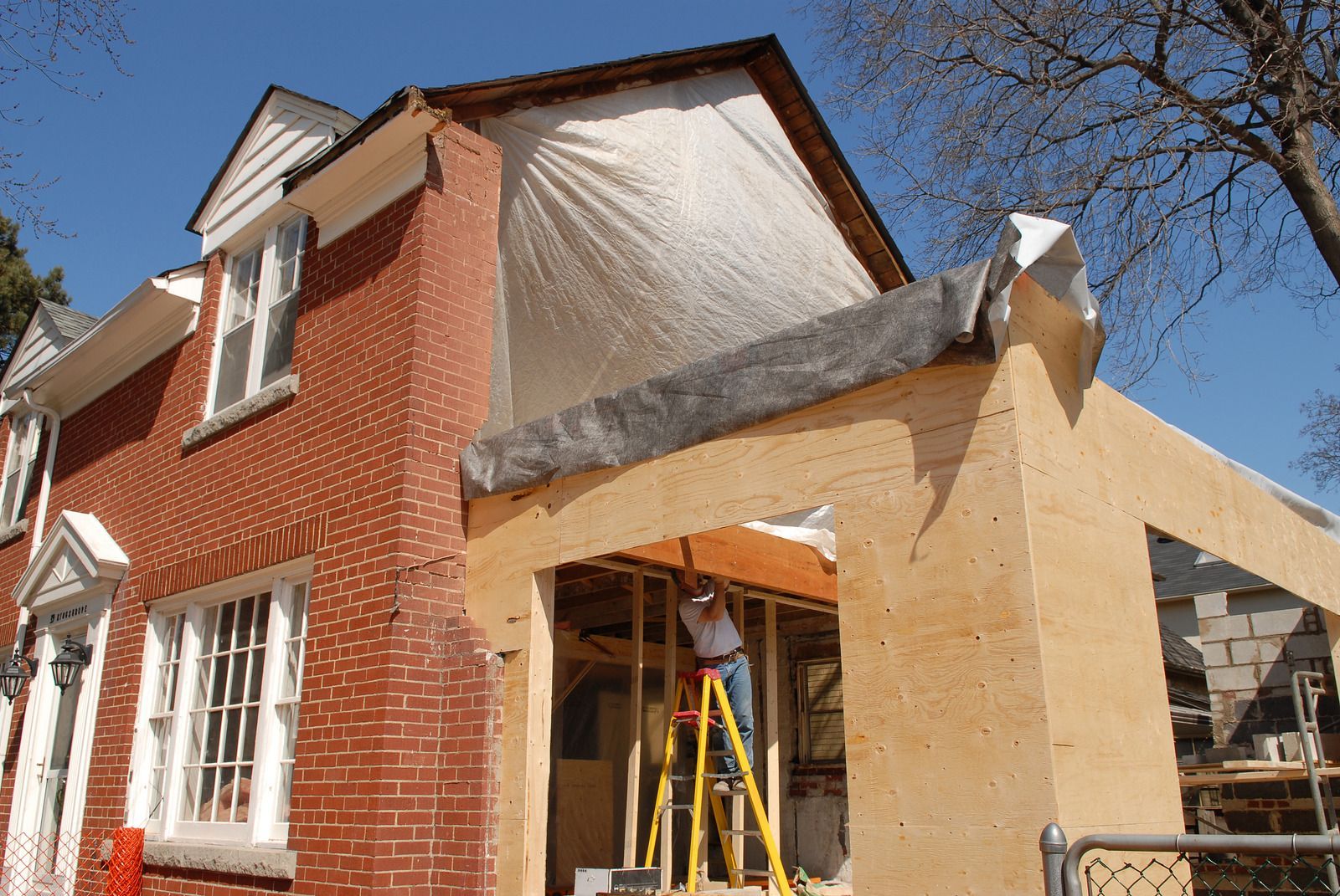 A brick house with a covered area under construction. A worker stands on a ladder inside the structure, under a blue sky.