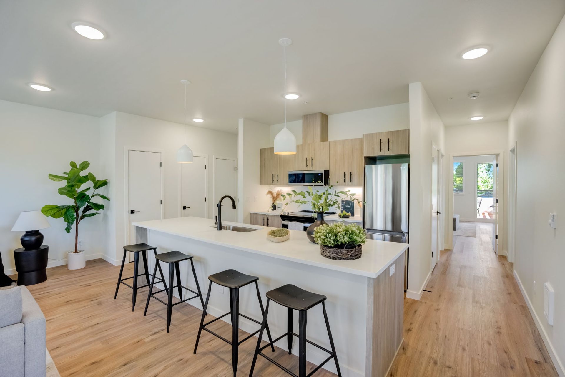 Modern kitchen with island, stools, and wooden cabinets. Hallway with natural wood floor, and entry door.