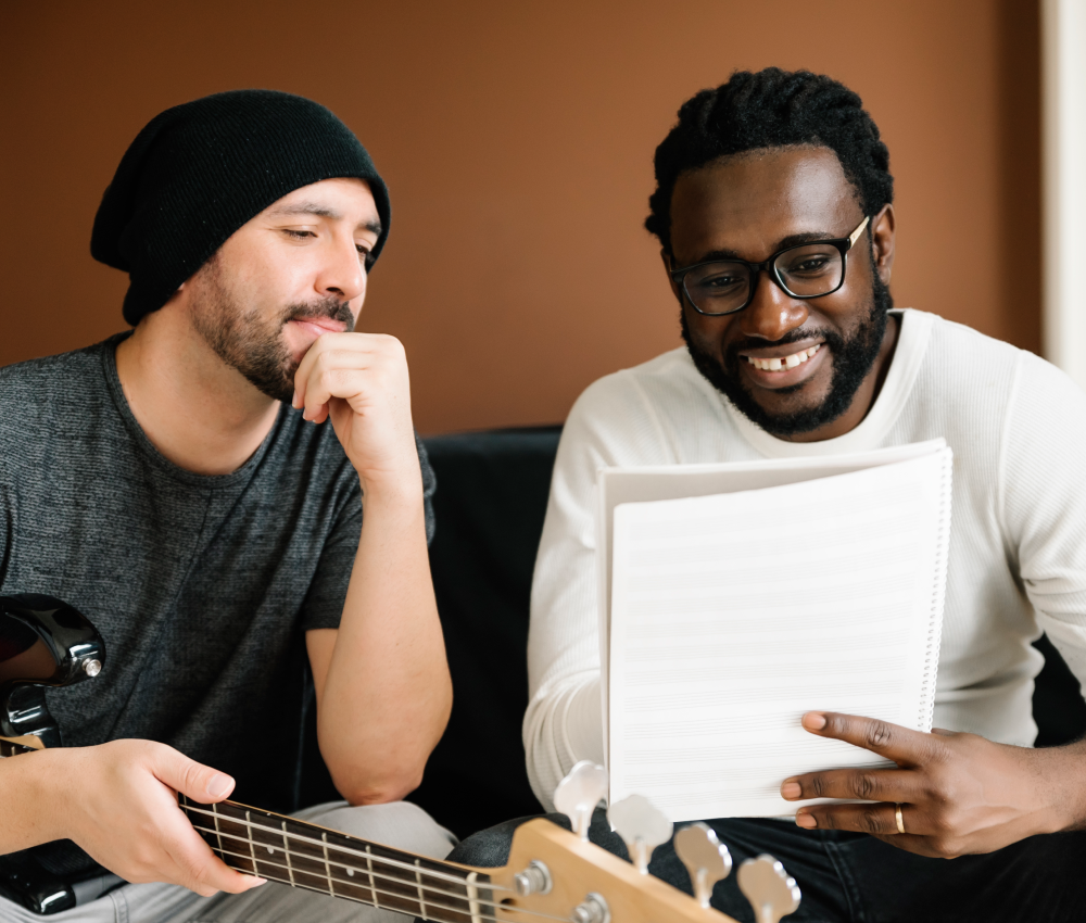 Two men are sitting on a couch one is playing a guitar and the other is reading a book.